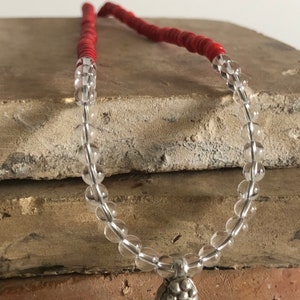 May include: A clear crystal beaded necklace with a silver pendant. The necklace is on a stack of brown tiles. The beads are round and the pendant is a cluster of small silver balls.