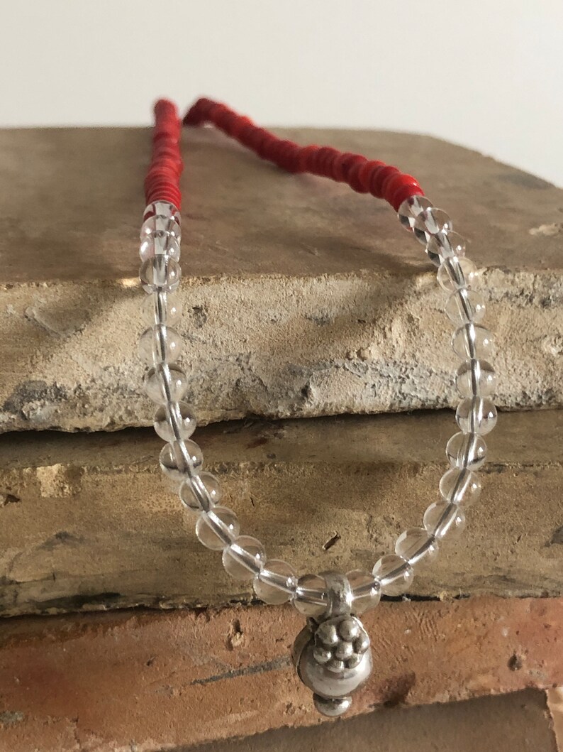 May include: A clear crystal beaded necklace with a silver pendant. The necklace is on a stack of brown tiles. The beads are round and the pendant is a cluster of small silver balls.