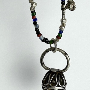 May include: A silver pendant necklace with a beaded chain. The chain features small, colorful beads in red, blue, green, and black. The pendant is a detailed, ornate silver sphere with a large silver ring above it.