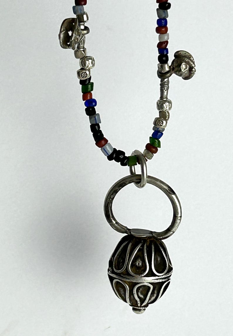 May include: A silver pendant necklace with a beaded chain. The chain features small, colorful beads in red, blue, green, and black. The pendant is a detailed, ornate silver sphere with a large silver ring above it.