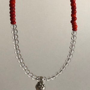 May include: A red and clear beaded necklace with a silver charm. The charm is a small, round object with a textured surface.