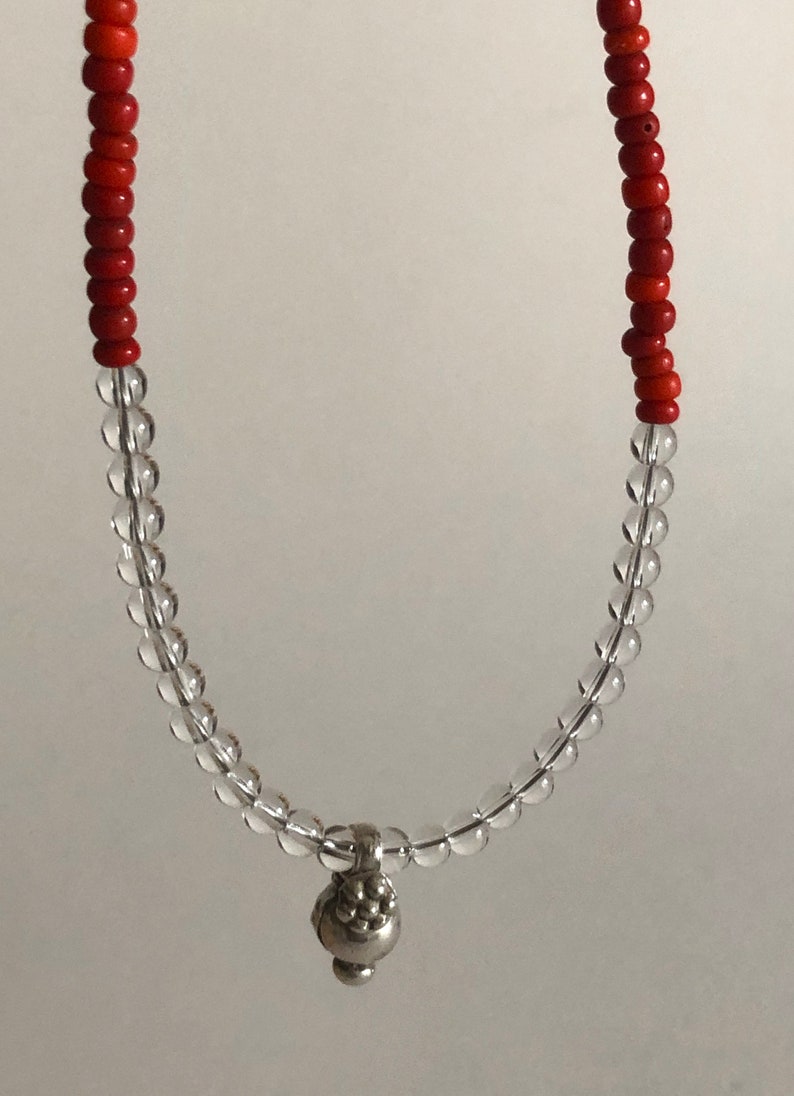May include: A red and clear beaded necklace with a silver charm. The charm is a small, round object with a textured surface.