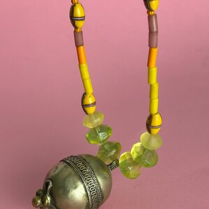 May include: A necklace featuring a large silver pendant with a decorative band and a small, ornate clasp. The necklace has yellow, orange, and brown beads, with green faceted beads near the pendant. The background is a solid pink.