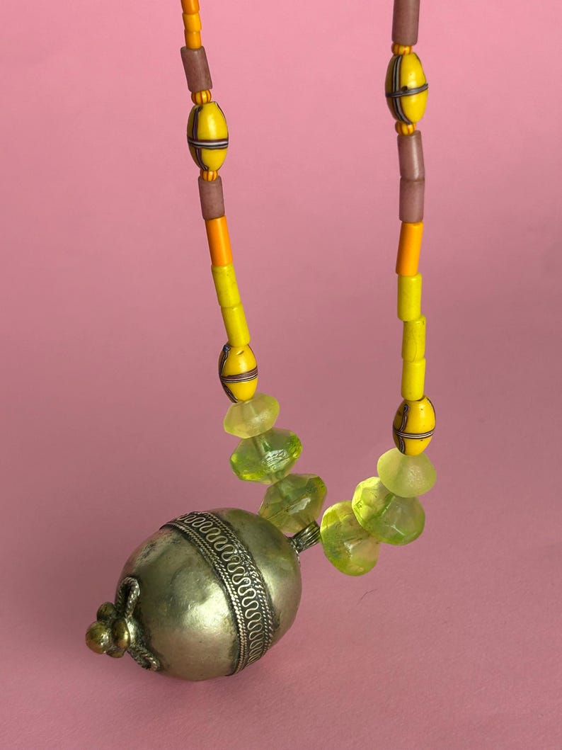 May include: A necklace featuring a large silver pendant with a decorative band and a small, ornate clasp. The necklace has yellow, orange, and brown beads, with green faceted beads near the pendant. The background is a solid pink.