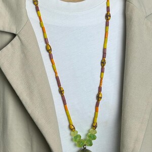 May include: A long beaded necklace with a silver pendant. The necklace features yellow, orange, and purple beads, with a cluster of green beads near the pendant. The pendant is a silver sphere with etched details.