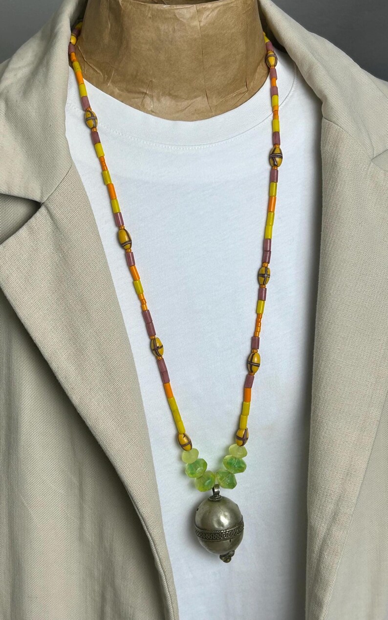 May include: A long beaded necklace with a silver pendant. The necklace features yellow, orange, and purple beads, with a cluster of green beads near the pendant. The pendant is a silver sphere with etched details.