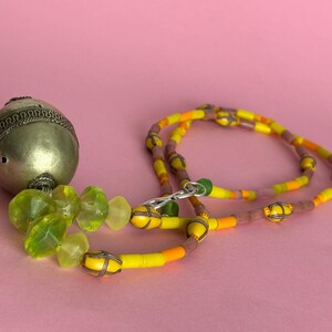 May include: A necklace with a silver metal pendant and a beaded chain. The pendant is a silver metal sphere with a decorative pattern. The chain features yellow, orange, and brown beads. The background is a solid pink.