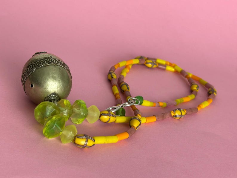 May include: A necklace with a silver metal pendant and a beaded chain. The pendant is a silver metal sphere with a decorative pattern. The chain features yellow, orange, and brown beads. The background is a solid pink.