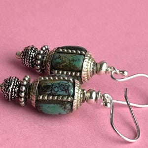 May include: A pair of handcrafted earrings with turquoise and silver-tone metal beads. The earrings feature a decorative design with a combination of turquoise stones and ornate silver-colored metalwork, suspended from silver-tone ear wires.