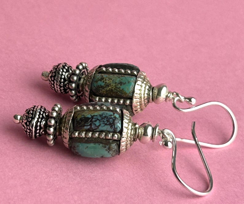 May include: A pair of handcrafted earrings with turquoise and silver-tone metal beads. The earrings feature a decorative design with a combination of turquoise stones and ornate silver-colored metalwork, suspended from silver-tone ear wires.