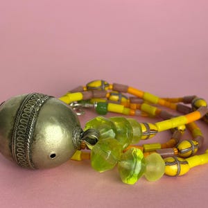 May include: A handcrafted necklace featuring a large silver-toned pendant with intricate detailing, complemented by strands of yellow, orange, and green beads. The necklace is set against a soft pink background.