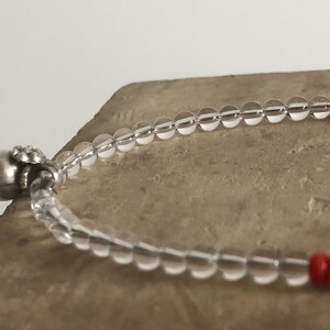 May include: A clear crystal beaded bracelet with a silver charm and red beads. The bracelet is on a light brown surface.