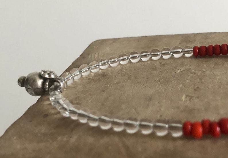 May include: A clear crystal beaded bracelet with a silver charm and red beads. The bracelet is on a light brown surface.