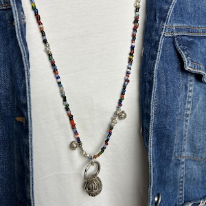 May include: A colorful beaded necklace with a silver pendant. The necklace features small, multi-colored beads and a silver pendant with a textured, spherical design. The necklace is worn over a white shirt and a denim jacket.