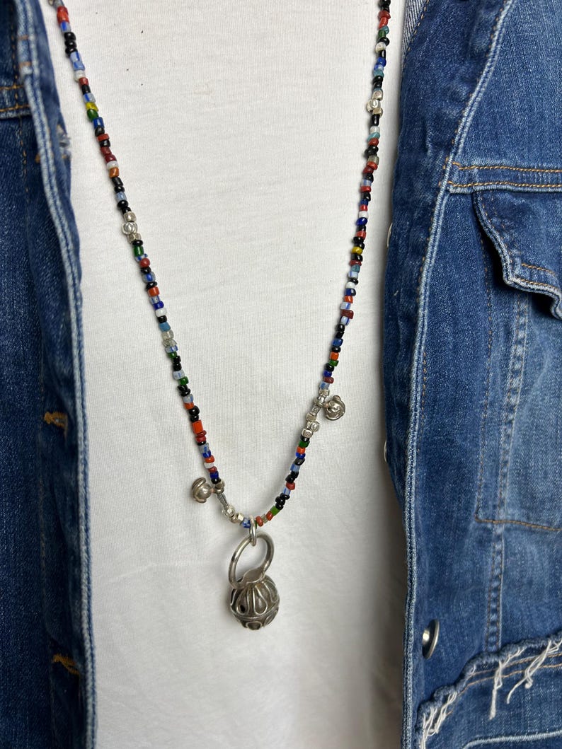May include: A colorful beaded necklace with a silver pendant. The necklace features small, multi-colored beads and a silver pendant with a textured, spherical design. The necklace is worn over a white shirt and a denim jacket.