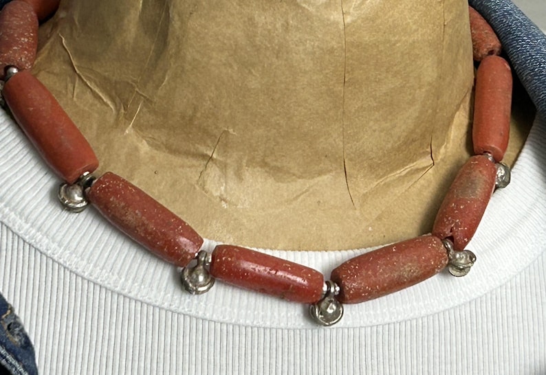 Pode incluir: Um colar com contas cil&iacute;ndricas castanho-avermelhadas, unidas com detalhes met&aacute;licos prateados. As contas t&ecirc;m uma apar&ecirc;ncia texturizada. O colar &eacute; exibido em uma camisa branca canelada.