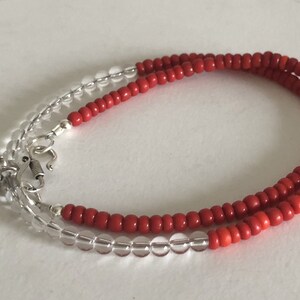 May include: A red and clear beaded bracelet with a silver charm. The bracelet is made of small, round beads and has a spring clasp.