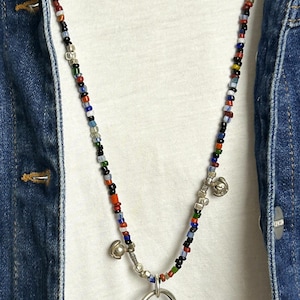 May include: A colorful beaded necklace with a silver pendant and small bells. The necklace features a variety of small, round beads in red, blue, green, and black. The pendant is a silver, ornate design. The necklace is worn over a white shirt and a denim jacket.