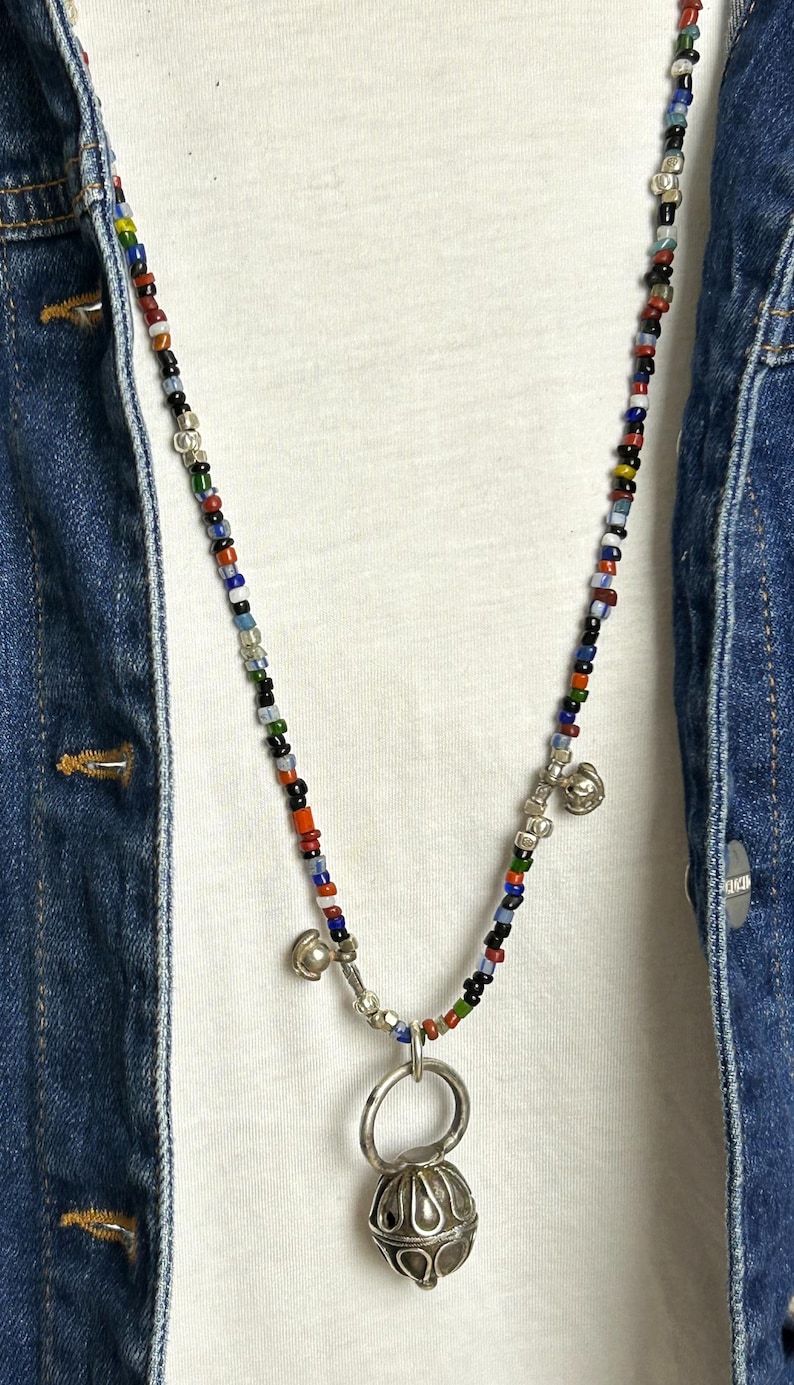May include: A colorful beaded necklace with a silver pendant and small bells. The necklace features a variety of small, round beads in red, blue, green, and black. The pendant is a silver, ornate design. The necklace is worn over a white shirt and a denim jacket.