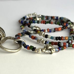 May include: A silver pendant with an ornate, textured design is connected to a beaded necklace. The necklace features small, colorful beads in various colors, including red, blue, and green. The jewelry is displayed on a white surface.
