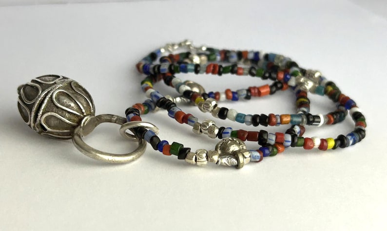 May include: A silver pendant with an ornate, textured design is connected to a beaded necklace. The necklace features small, colorful beads in various colors, including red, blue, and green. The jewelry is displayed on a white surface.