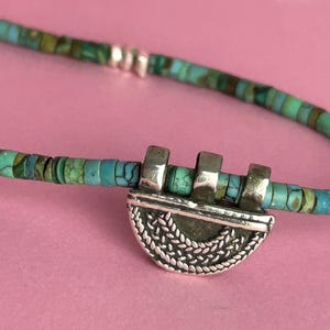 Puede incluir: Collar turquesa y plata. El collar presenta pequeñas cuentas redondas de turquesa en tonos azules y verdes. Un colgante plateado con forma de media luna y diseño trenzado es el punto focal.
