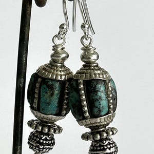 May include: A pair of silver earrings with turquoise-colored, oval-shaped beads. The earrings have intricate silver detailing and a decorative, beaded bottom. The ear wires are silver.