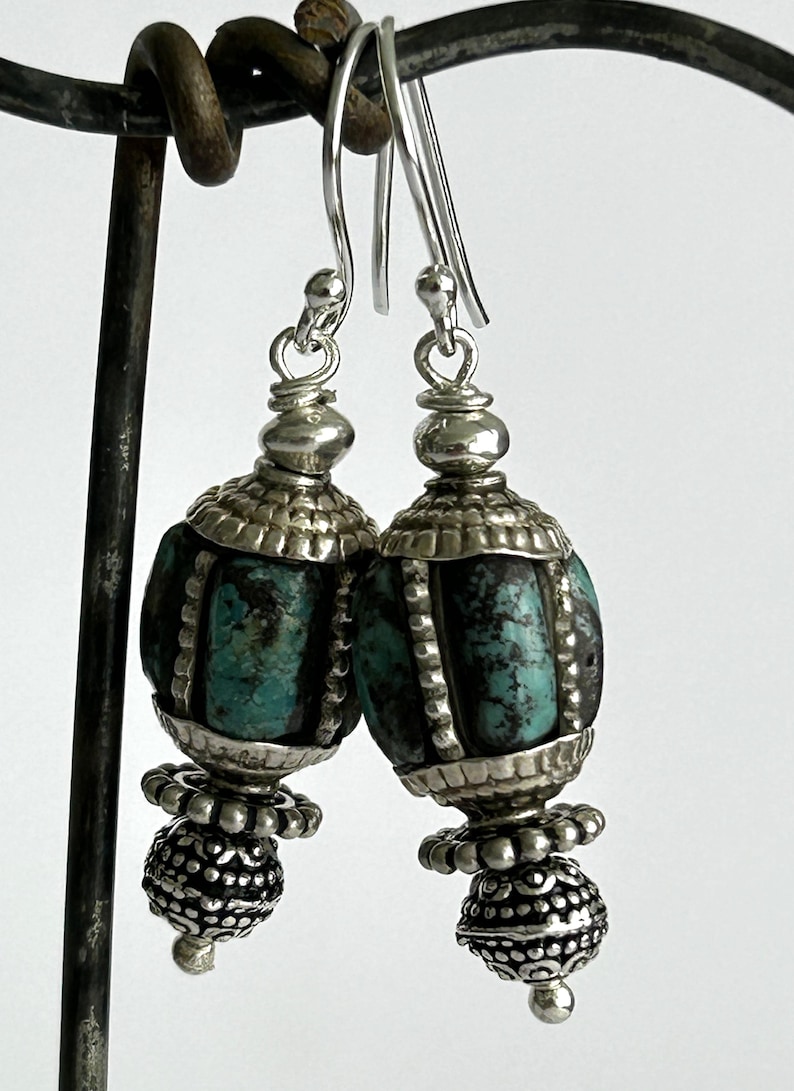 May include: A pair of silver earrings with turquoise-colored, oval-shaped beads. The earrings have intricate silver detailing and a decorative, beaded bottom. The ear wires are silver.