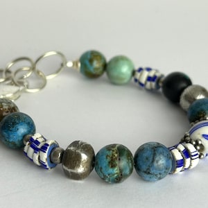 May include: A colourful beaded bracelet with a mix of beads. The bracelet has blue, green, and silver beads, plus white and blue striped beads. It is fastened with a silver clasp.