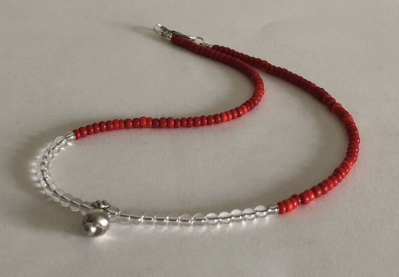 May include: A red and clear beaded necklace with a silver charm. The necklace is made of small, round beads and has a simple design.