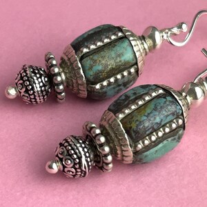 May include: A pair of handcrafted earrings featuring turquoise-colored beads with silver-toned accents. The earrings have a unique design with intricate detailing and a hook closure. The background is a solid pink color.