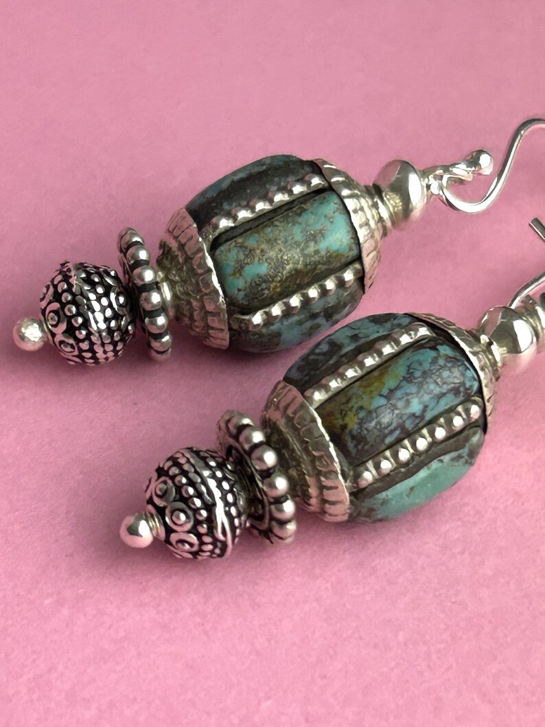 May include: A pair of handcrafted earrings featuring turquoise-colored beads with silver-toned accents. The earrings have a unique design with intricate detailing and a hook closure. The background is a solid pink color.