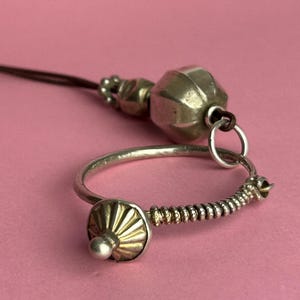 May include: A silver ring with a decorative gold and silver bead, connected to a silver and gold beaded spiral and a silver and gold decorative bead. A brown cord is attached to a silver bead and a larger silver bead.