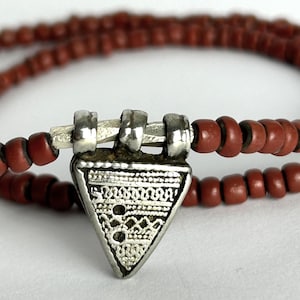 Puede incluir: Un collar con un colgante triangular de plata con intrincados diseños, suspendido de una cadena de pequeñas cuentas de color marrón rojizo. La joya se muestra sobre un fondo blanco, resaltando los detalles.