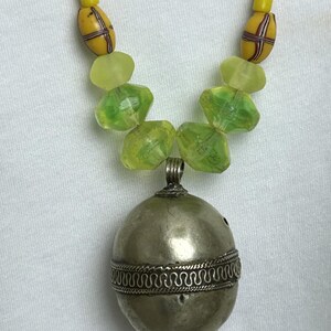May include: A handcrafted necklace featuring a silver-toned pendant with intricate detailing. The necklace includes yellow and green beads, creating a unique and eye-catching design. The pendant is approximately 1.5 inches in diameter.