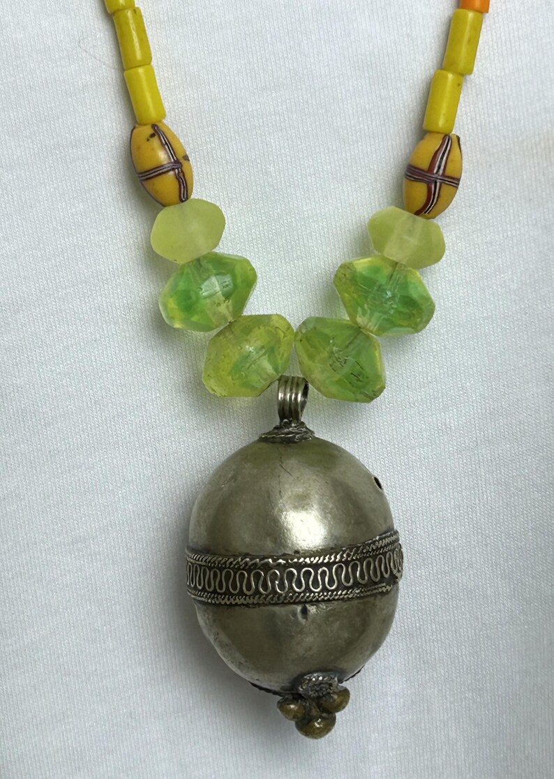 May include: A handcrafted necklace featuring a silver-toned pendant with intricate detailing. The necklace includes yellow and green beads, creating a unique and eye-catching design. The pendant is approximately 1.5 inches in diameter.