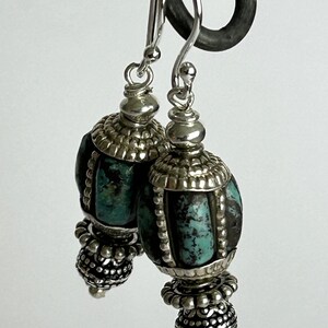 May include: A pair of silver dangle earrings with turquoise-colored stone beads. The earrings have a detailed design with silver caps and decorative elements. The turquoise beads are oval-shaped and have a textured appearance. The earrings are suspended from silver hooks.