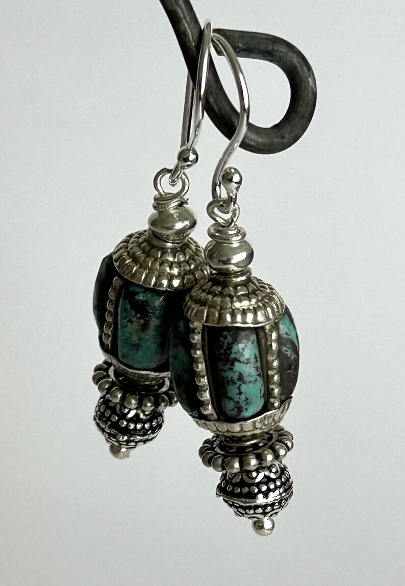 May include: A pair of silver dangle earrings with turquoise-colored stone beads. The earrings have a detailed design with silver caps and decorative elements. The turquoise beads are oval-shaped and have a textured appearance. The earrings are suspended from silver hooks.