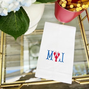May include: White hand towel with blue embroidered letters "M" and "J" with a red lobster in between the letters.