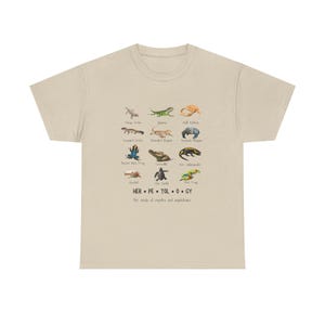 May include: Beige t-shirt featuring a collection of reptile and amphibian illustrations. The shirt includes the text "HER.PE.TOL.O.GY" and the phrase "The study of reptiles and amphibians."