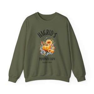 May include: Olive green sweatshirt with the text "Hagrid's Pumpkin Farm" above a watercolor illustration of a pumpkin and flowers. The words "Magically Grown" are below the illustration. The sweatshirt has long sleeves and a crew neck.