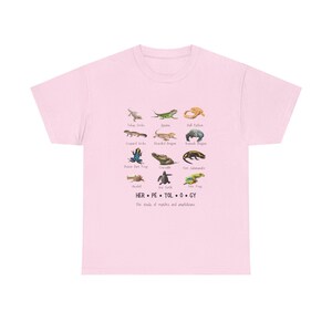 May include: Light pink t-shirt featuring a colorful illustration of various reptiles and amphibians, including geckos, lizards, and frogs. The word "HERPETOLOGY" is printed below the illustrations, along with the phrase "The study of reptiles and amphibians."