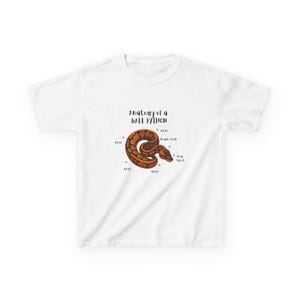 Peut inclure: T-shirt blanc avec une illustration détaillée d'un python royal et des étiquettes anatomiques. Le serpent est brun et beige, avec le texte "Anatomy of a Ball Python" au-dessus de l'illustration. Le motif est centré sur le t-shirt.