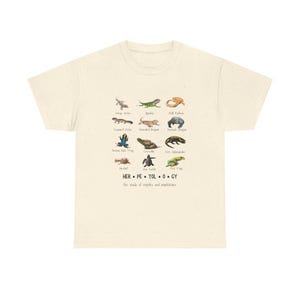 May include: Cream-colored t-shirt featuring a colorful illustration of various reptiles and amphibians, including geckos, iguanas, and frogs. The word "HERPETOLOGY" is printed below the illustrations, along with the phrase "The study of reptiles and amphibians."