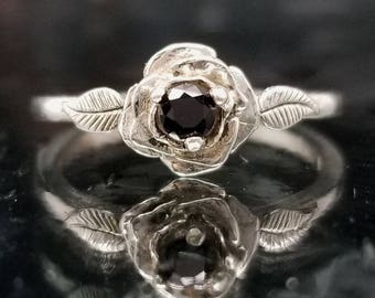 Puede incluir: Un anillo de plata con una piedra preciosa negra engastada en un diseño de rosa. El anillo tiene hojas delicadas a cada lado de la rosa.