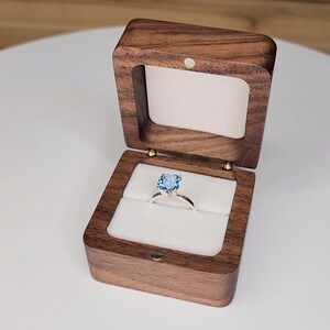 Puede incluir: Una caja de anillo de madera con una tapa con bisagras, abierta para revelar un anillo de piedra azul sobre un cojín blanco.