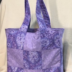 May include: A purple quilted tote bag with a patchwork design. The bag has two handles and is made of a soft, fabric material.
