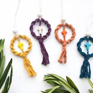 Może przedstawiać: Cztery makramowe ozdoby w kształcie krzyża w kolorze żółtym, fioletowym, pomarańczowym i turkusowym. Każda ma pasującą frędzlę i krzyż w kontrastującym kolorze. Ozdoby zawieszone są na białym sznurku z białą koralikiem na górze. Zielone liście w tle.