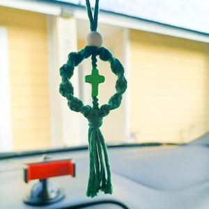 Może przedstawiać: Zielony urok do samochodu z makramy z okrągłym wzorem i zielonym krzyżem pośrodku. Urok jest zawieszony na zielonym sznurku i drewnianej koraliku. Urok wisi przed deską rozdzielczą samochodu.