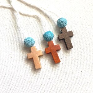May include: Three wooden cross pendants in shades of brown, orange, and tan, each with a light blue, porous bead. The crosses hang on white cords, against a textured white background. The beads are approximately 1 cm in diameter.